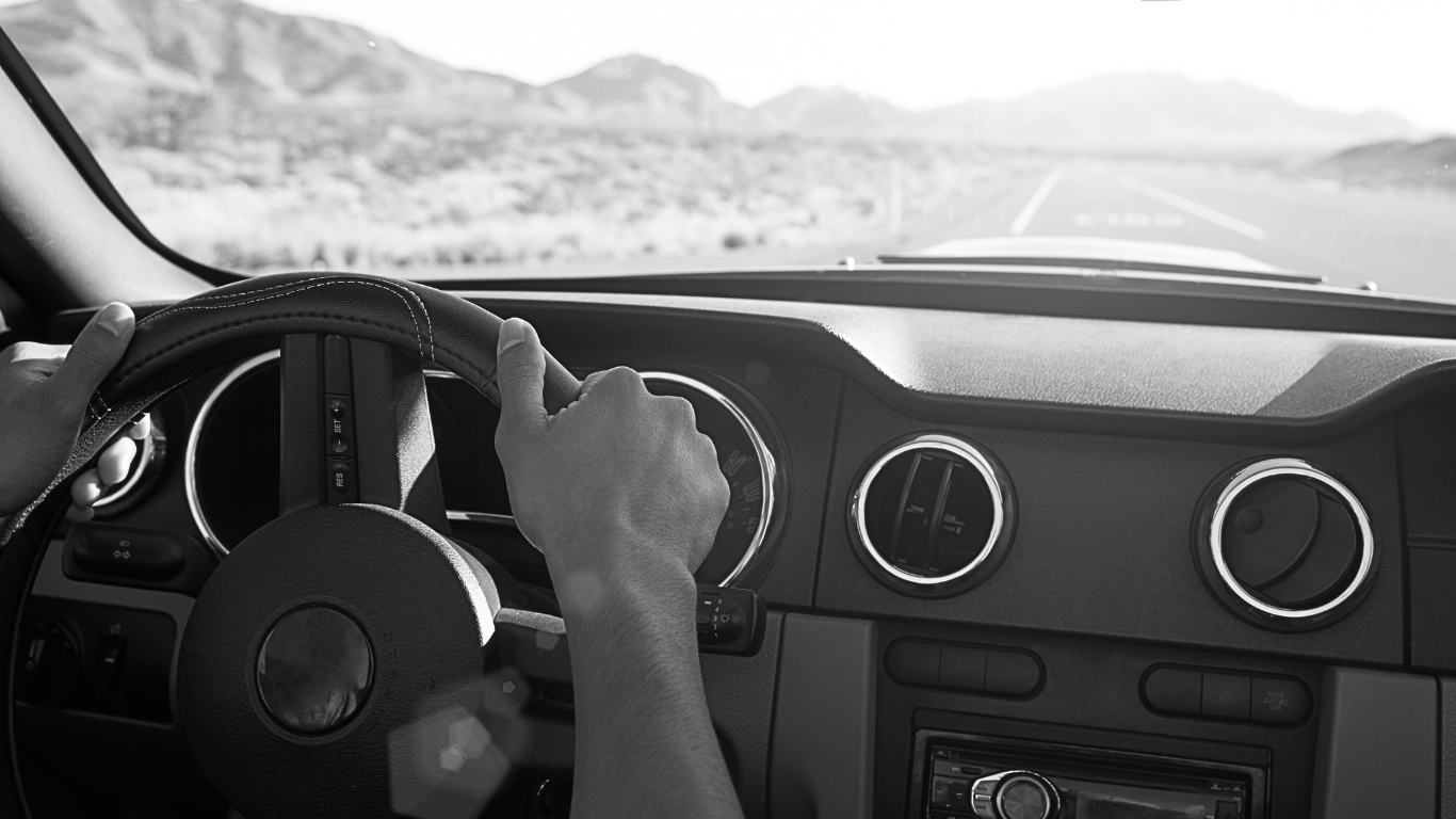 Black and white stock image of person driving left hand drive car - can see steering wheel and dashboard.